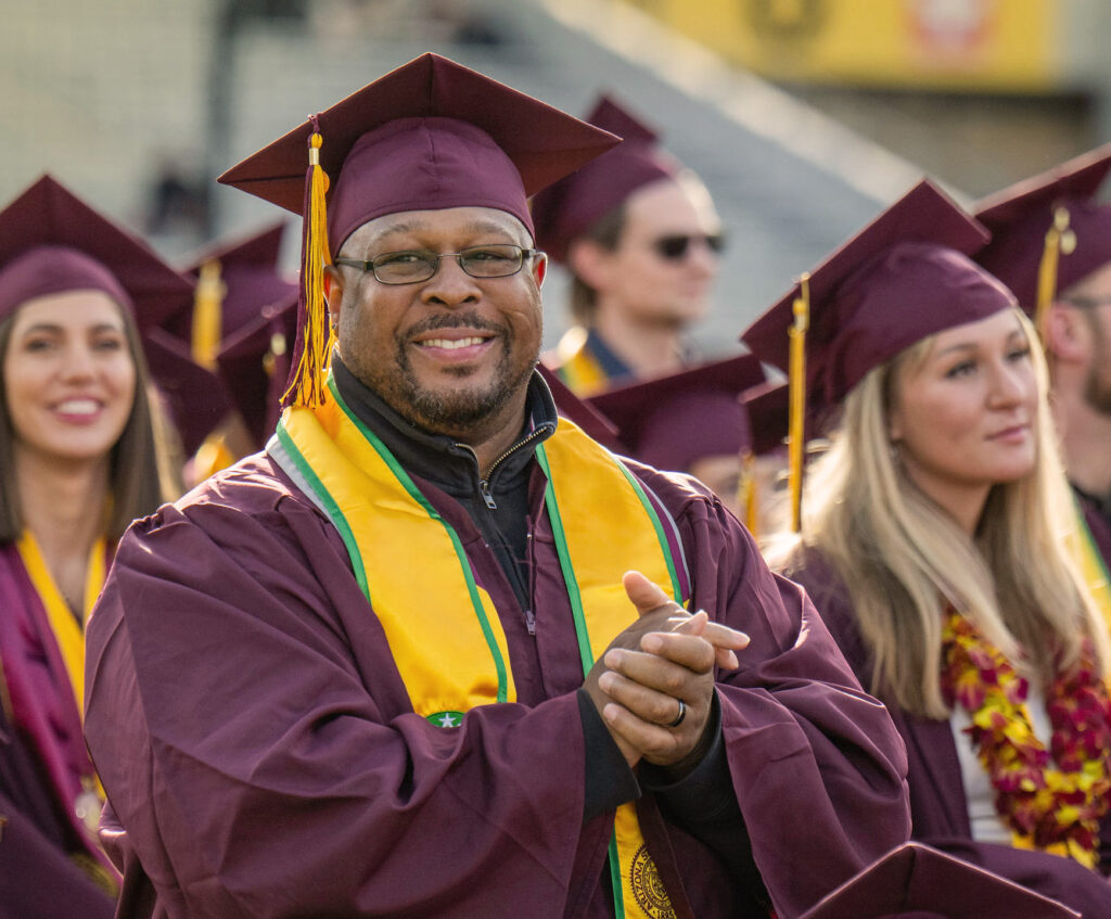 A person wearing an ASU cap and gown and yellow sash stands cheering during a commencement ceremony.