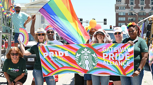 Starbucks partners stand behind a rainbow sign that reads, "Starbucks Pride Network". One person is holding a rainbow flag. 
