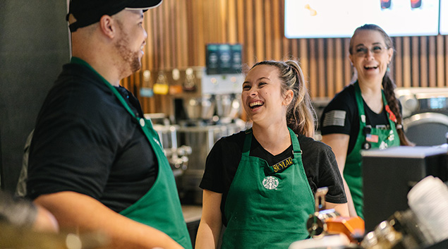 Three individuals wearing green aprons smile and laugh as they work together at a Starbucks