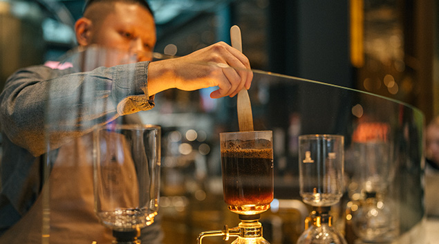 An individual standing at a coffee siphon, carefully stirring the contents with a long-handled spoon.