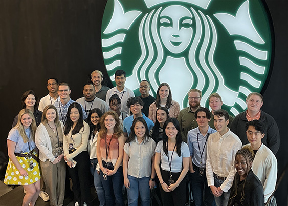 A group of students stand in front of the Starbucks Siren sign at the Seattle Headquarters. 