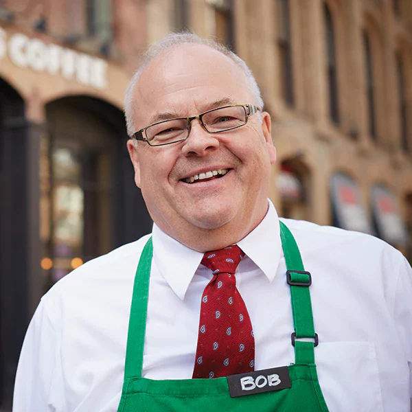 A person wearing a green apron, white button down shirt and red tie smiles.