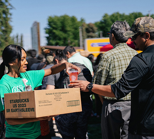 A person hands a cold Starbucks beverage to another person standing in line at a community service event.
