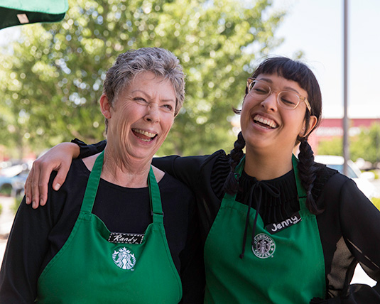 Two individuals, wearing Starbucks green aprons, share a warm embrace, laughing joyfully.