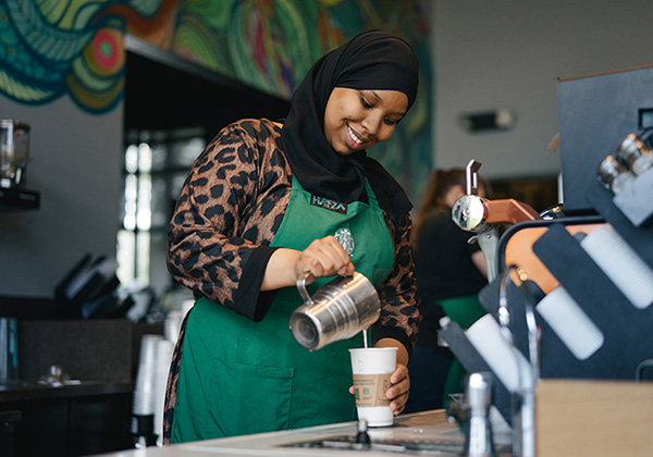 A person wearing a hijab and a Starbucks green apron smiles while preparing a handcrafted coffee beverage.