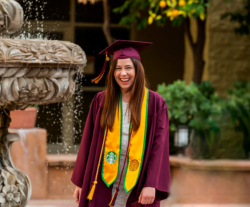 A person wearing a maroon ASU cap and gown adorned with a yellow sash, displaying both the University of Arizona and Starbucks logos, smiles.