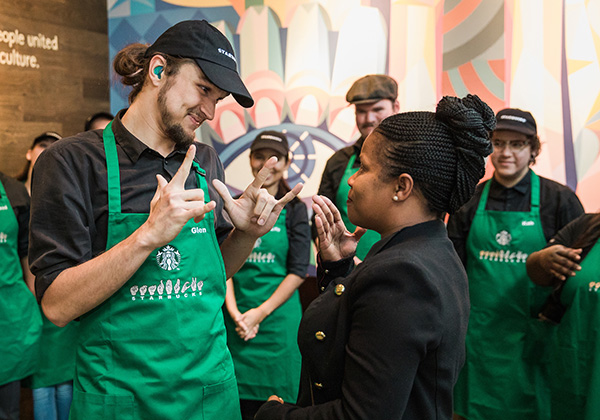 Two individuals wearing Starbucks green aprons communicate with each other using sign language, surrounded by fellow employees.