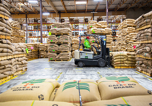 A person operating a forklift in a warehouse filled with neatly stacked burlap coffee bags, maneuvers through the warehouse