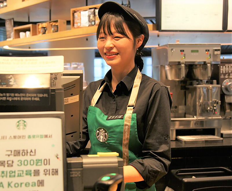 A person wearing a Starbucks green apron and a black hat smiles warmly while standing behind a register.