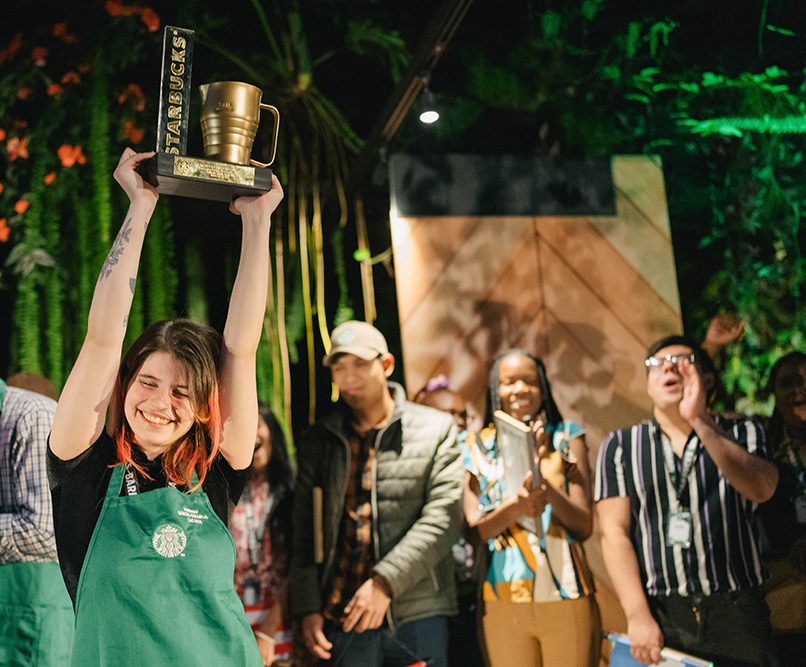 A person wearing a Starbucks green apron smiles while holding up a trophy in celebration.
