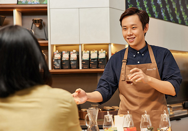 A person wearing a tan apron smiles while engaging with a customer and presenting a flight of drinks at a Shanghai Starbucks cafe.