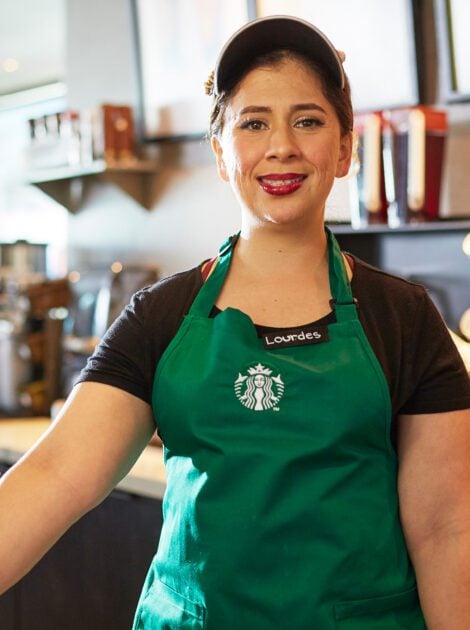 A person wearing a green apron and Starbucks visor smiles at the camera.