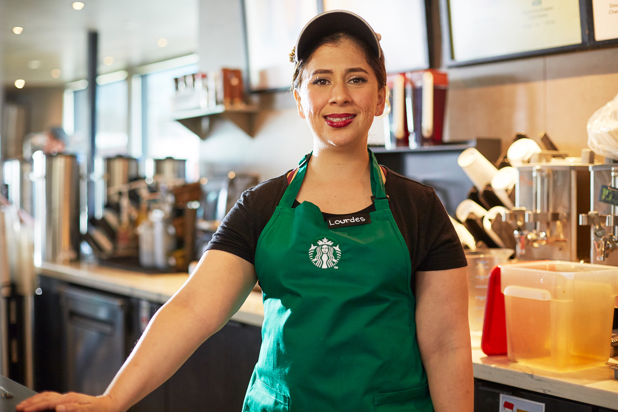 A person wearing a green apron and Starbucks visor smiles at the camera.
