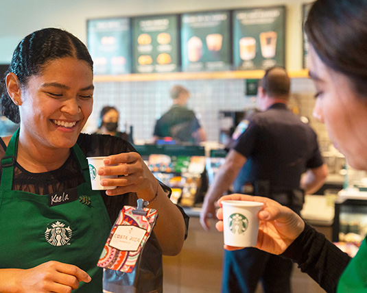 Two people wearing green Starbucks aprons sit together, sharing coffee and smiling. A bag of Costa Rica Reserve coffee sits on the table between them.
