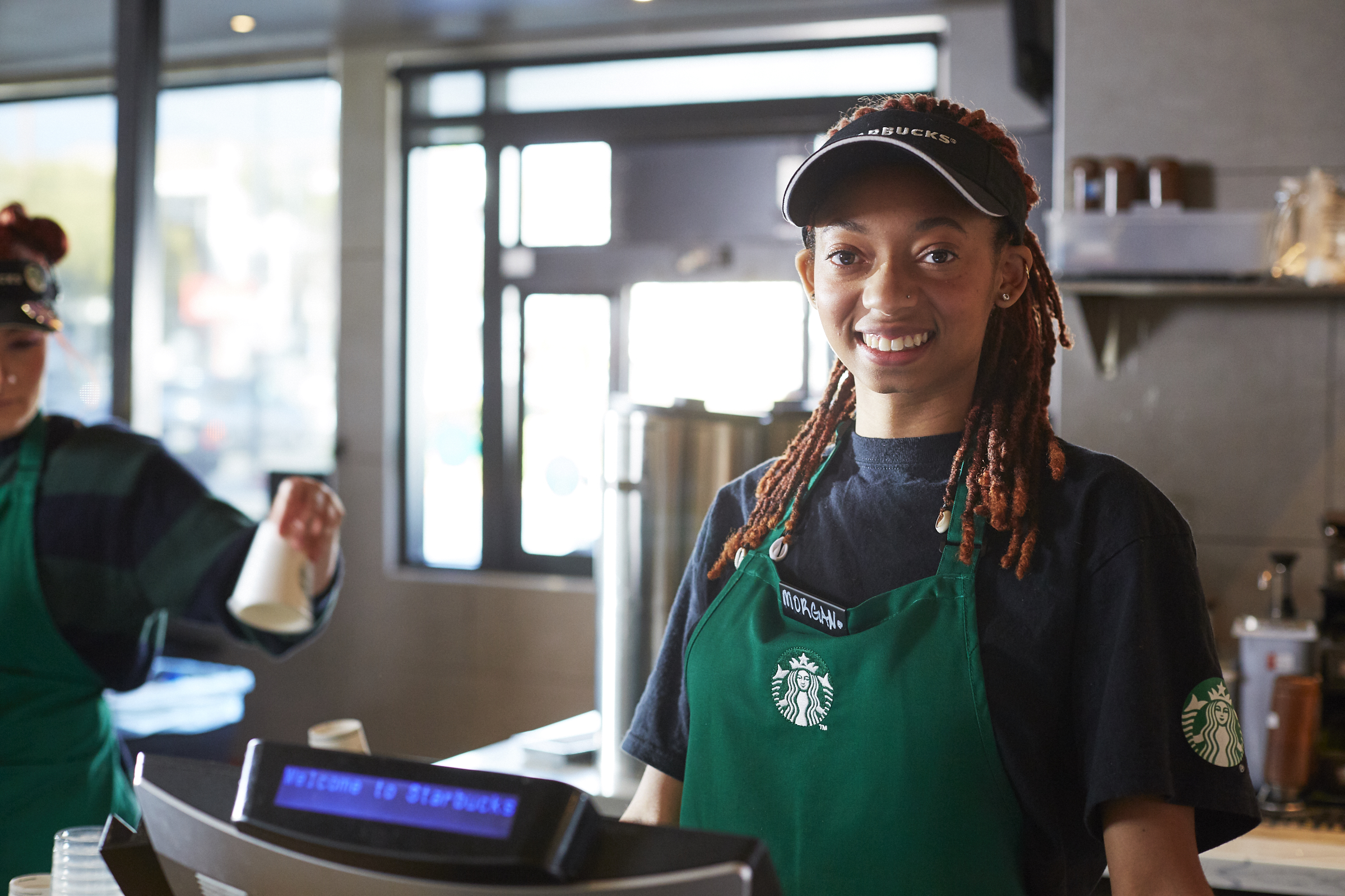 A Starbucks employee behind the counter in a store smiles while wearing a green apron and Starbucks visor.