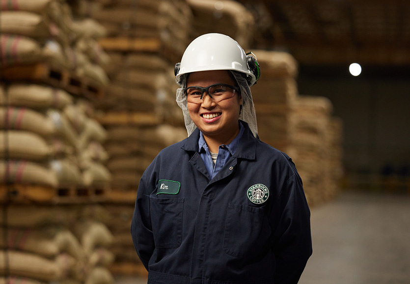 A person wearing coveralls with a Starbucks logo smiles, they are wearing a white hard hat in a warehouse.