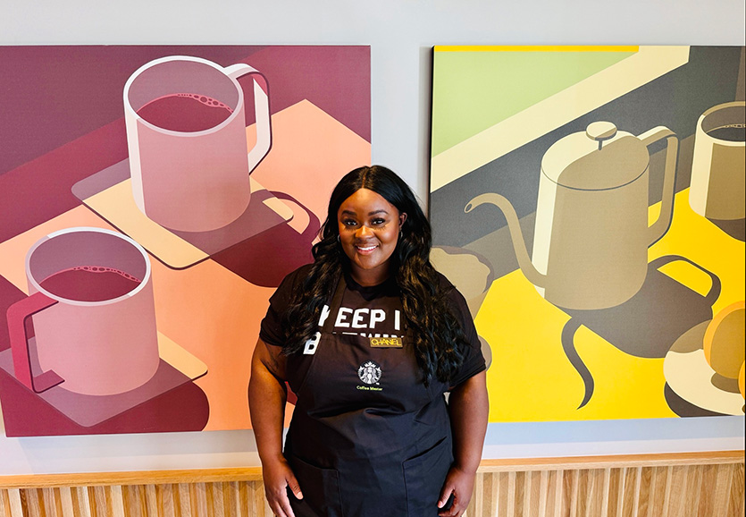 A person stands in front of an artistic drawing of coffee cups wearing a black apron.