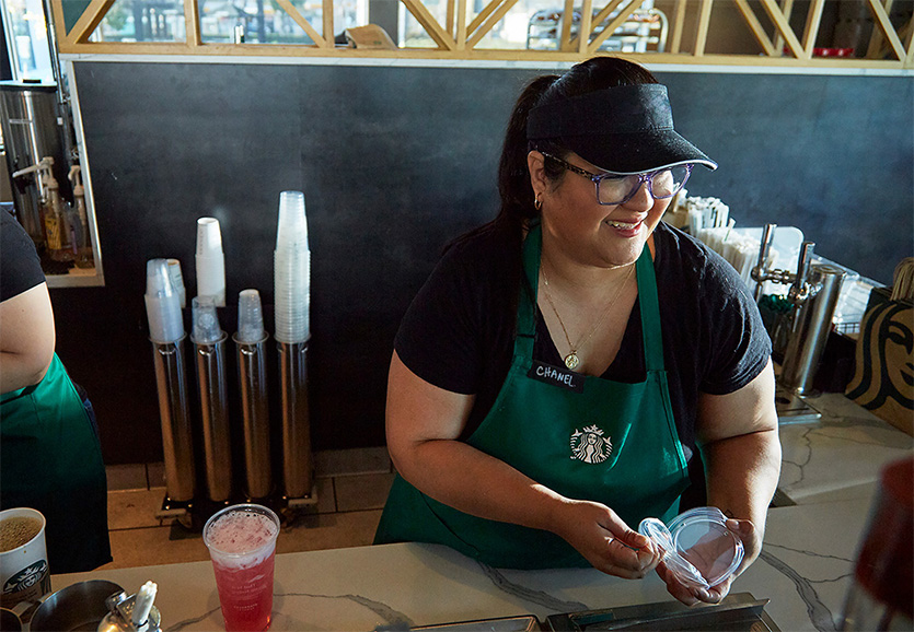 A person wearing a Starbucks green apron and visor smiles while handing off a cold beverage lid.