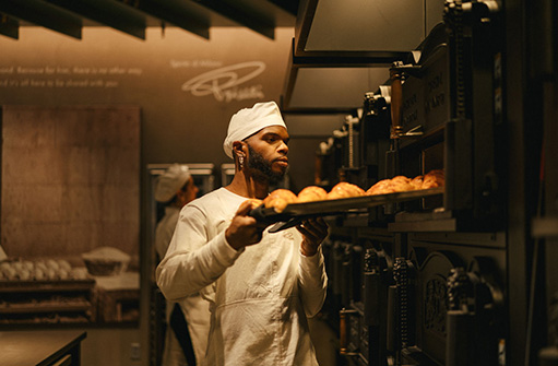 A person dressed in a baker's uniform loads a pan of bread into an oven.