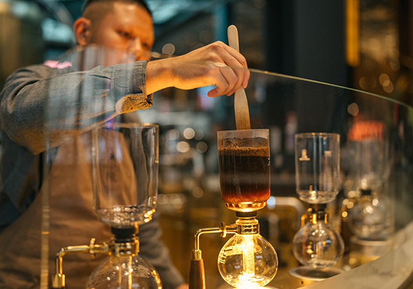 An individual standing at a coffee siphon, carefully stirring the contents with a long-handled spoon.