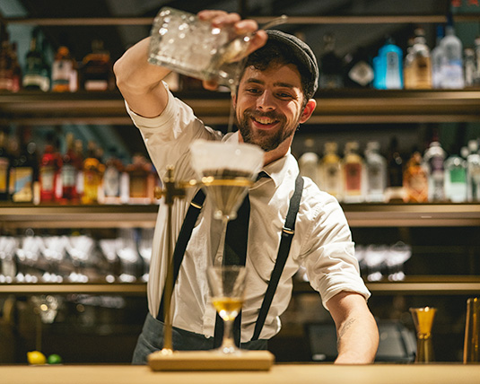 A person stands behind a bar, smiling as they mix a cocktail through a siphon.