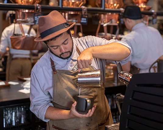 A person wearing a tan apron and brown hat concentrates on crafting a latte, pouring milk into the coffee.