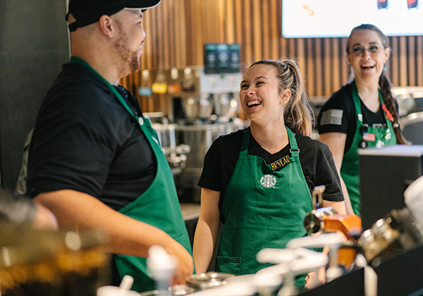 Three individuals wearing green aprons smile and laugh as they work together at a Starbucks 
