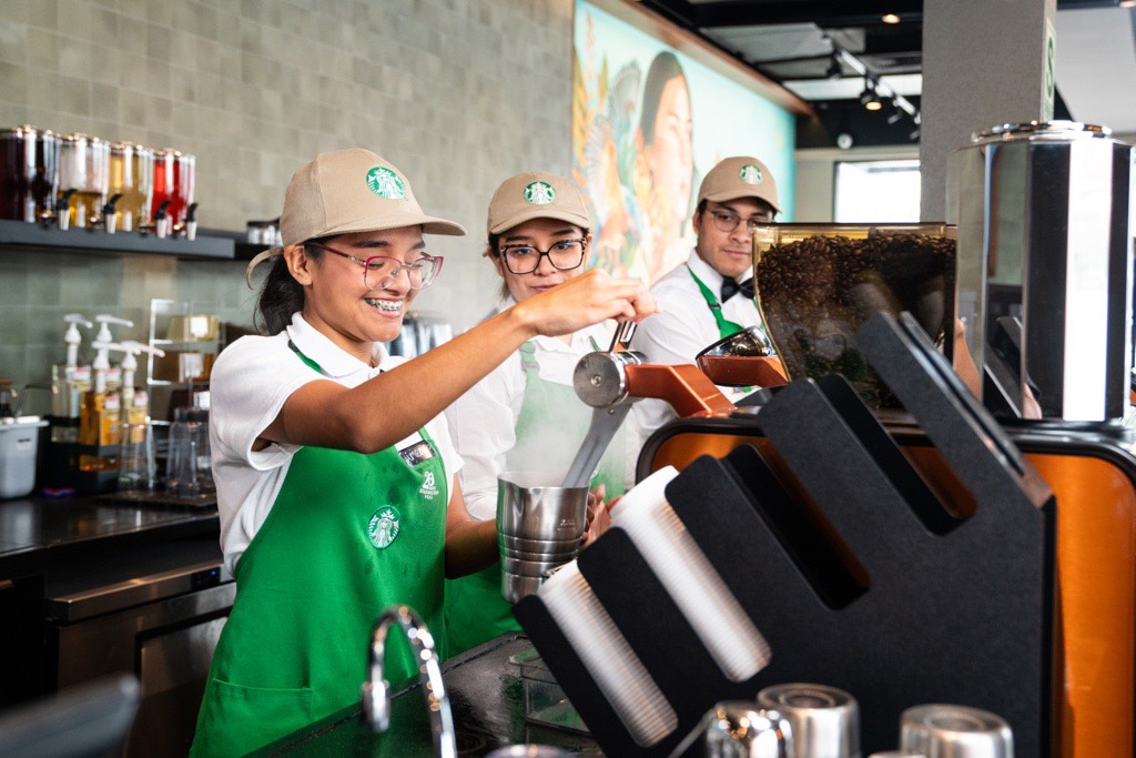 A person, wearing a Starbucks green apron and a tan Starbucks hat, smiles while preparing an espresso beverage. They stand alongside two coworkers who are dressed similarly.