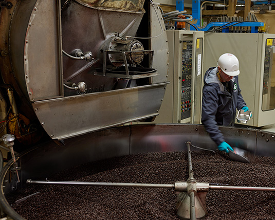 A person wearing a hardhat stands in a manufacturing warehouse, observing coffee beans in a large coffee roaster.