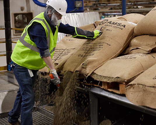 A person wearing a hardhat and a bright green vest stands in front of a cut-open burlap sack of coffee, emptying its contents.