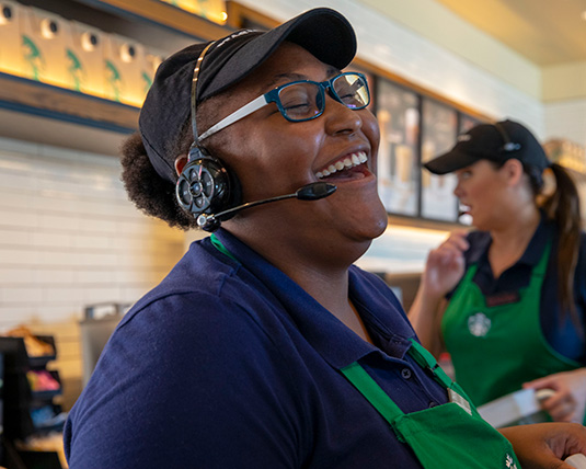 A person wearing a green apron and drive-thru headpiece smiles while working.