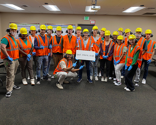 A large group of people, dressed in orange vests and yellow hardhats, pose in front of a camera while holding a sign that says 'Best Interns Ever'.
