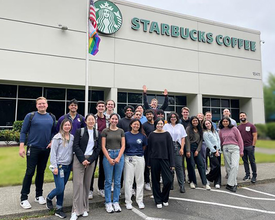 A group of individuals of diverse backgrounds stand in front of a building with a Starbucks sign.