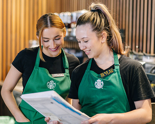 Two individuals wearing green aprons smiling as they review a document together.