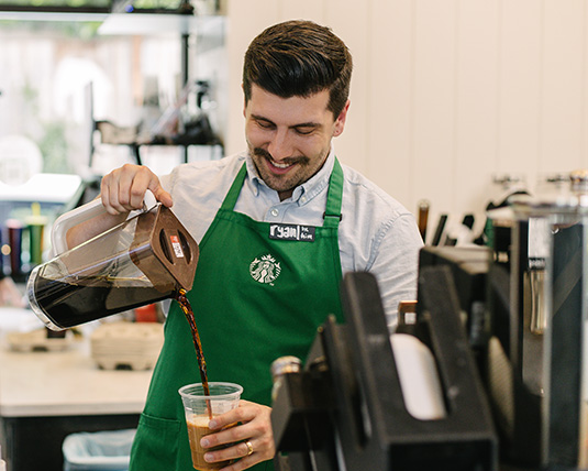 A person in a green Starbucks apron pours coffee in a store.