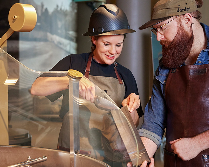 Two people are standing at a coffee roaster scooping beans into a coffee bag.