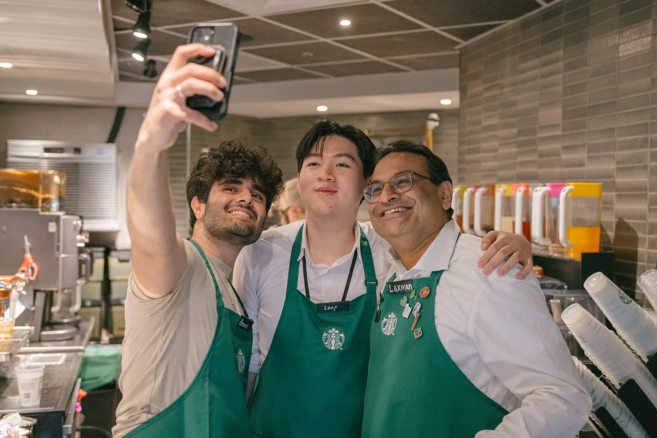 Two people in green aprons pose with Starbucks CEO Laxman Narasimhan behind a counter, all smiling as they take a selfie with a smartphone.