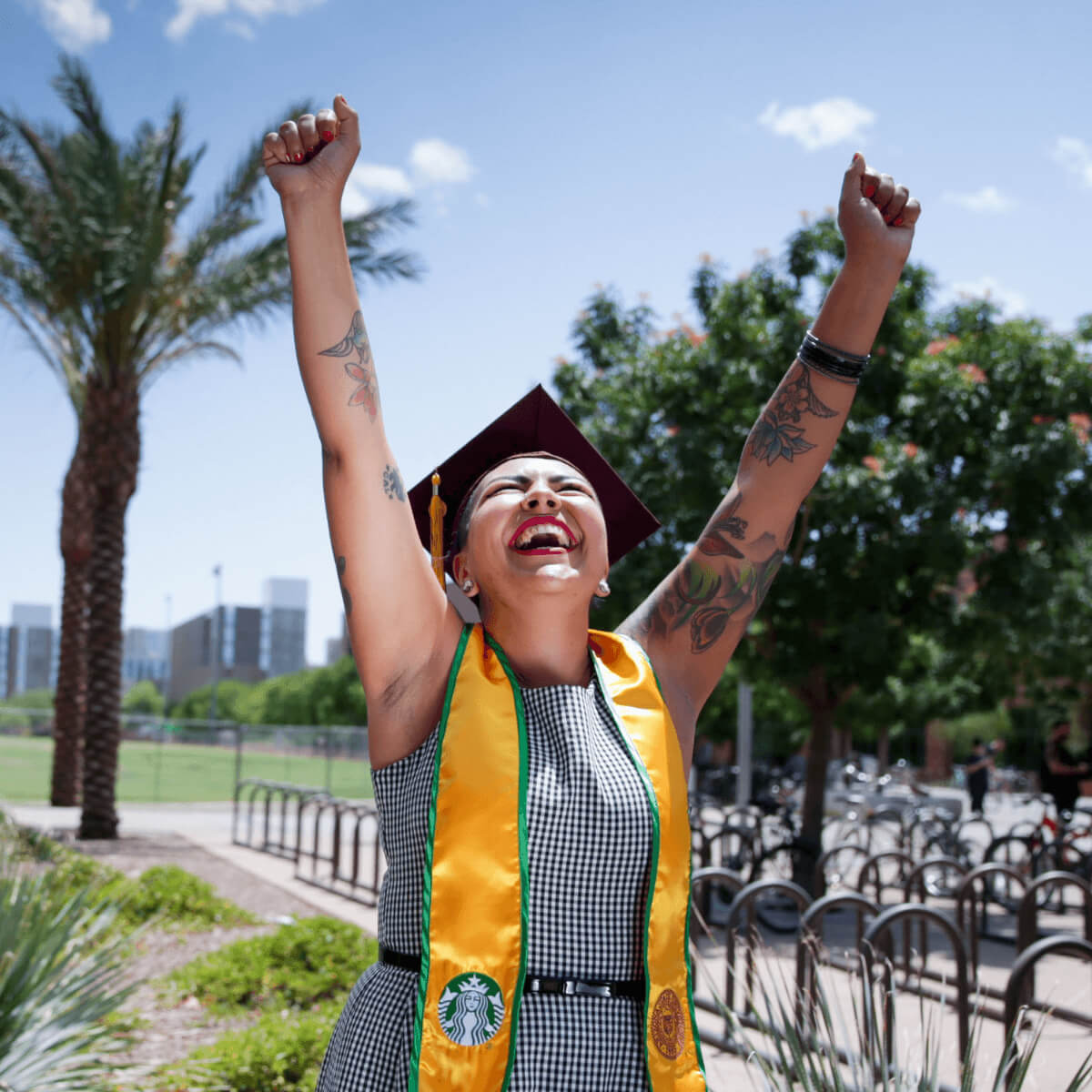 A person wearing a maroon ASU cap and gown adorned with a yellow sash, displaying both the University of Arizona and Starbucks logos, smiles.