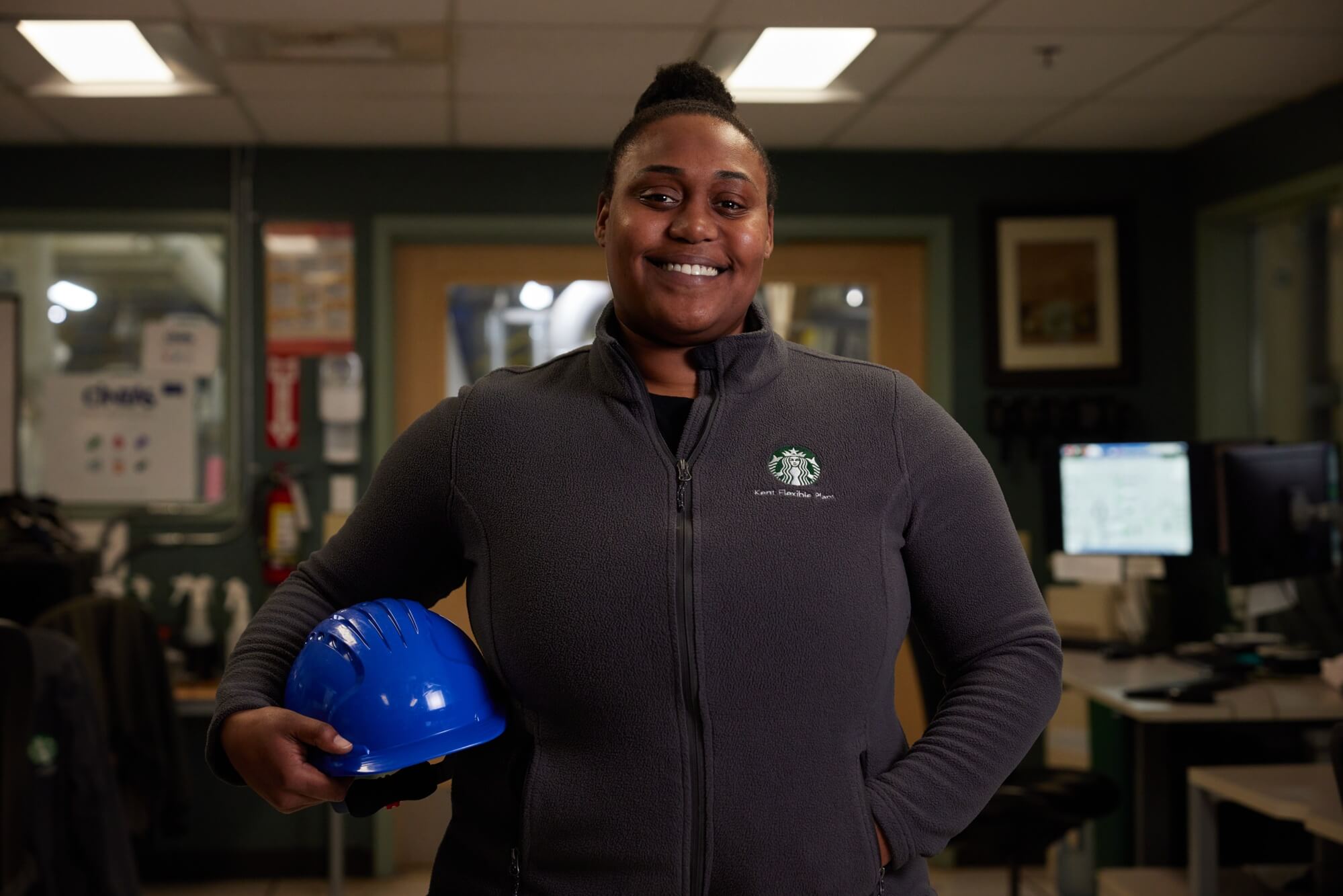 A person wearing a Starbucks branded zip-up jacket holds a blue hard hat and smiles .