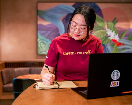 A Starbucks employee sits at a table inside a Starbucks coffeehouse and works on her computer. She is a student in the SCAP program and wears a shirt reading, “Coffee and College”.