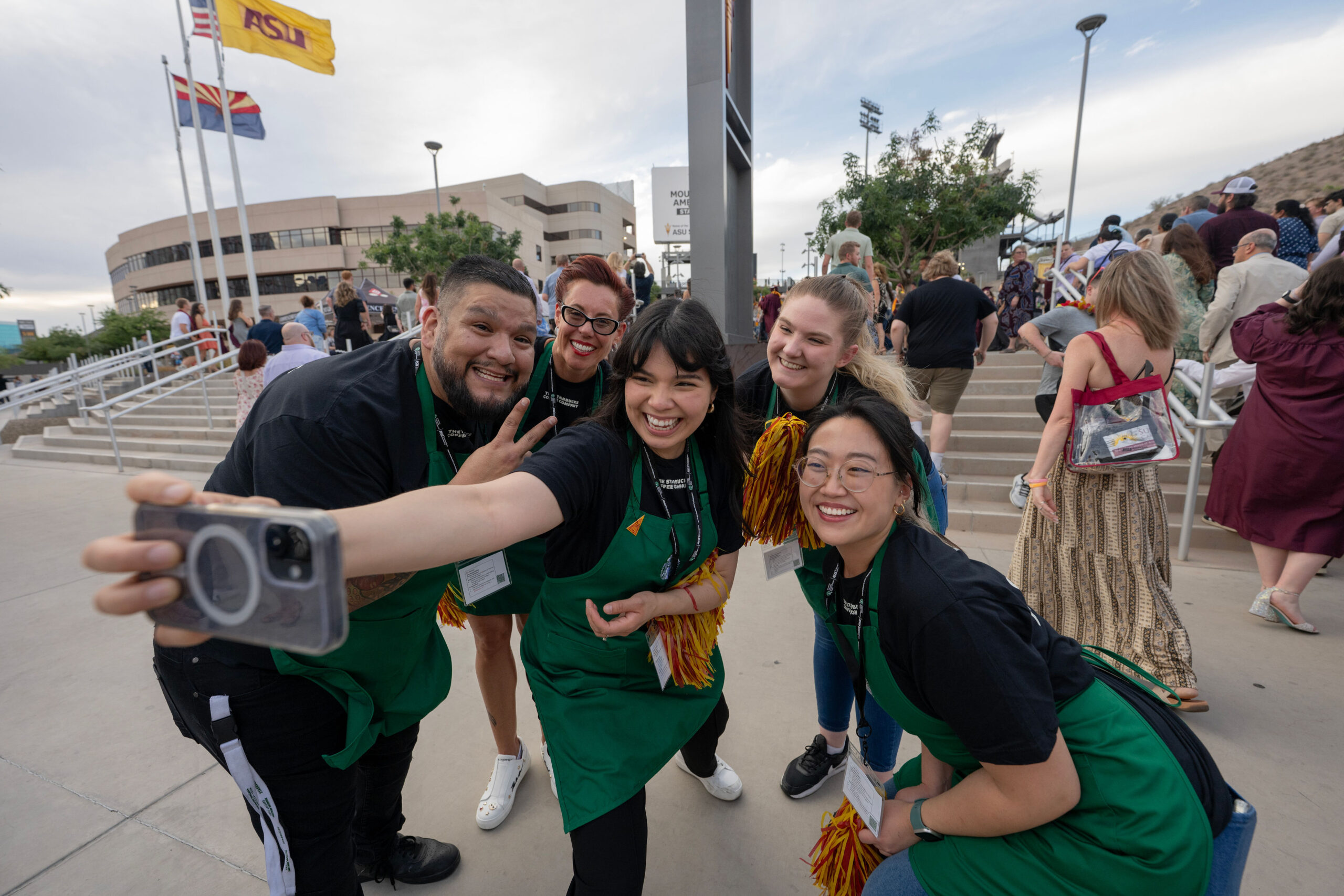 A group of Starbucks employees wearing green aprons stand together for a selfie photo moment.