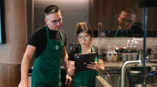 Two Starbucks employees stand next to each other in a coffeehouse and look quizzically at a handheld device.