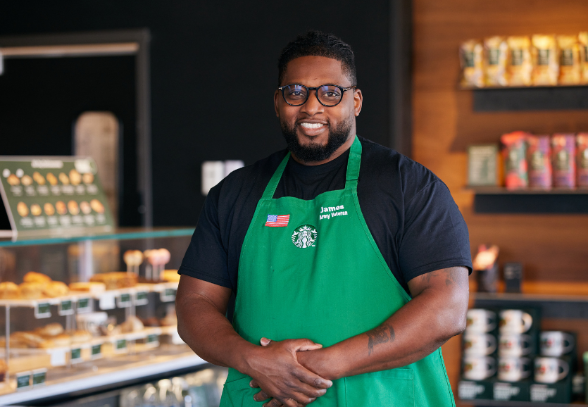A Starbucks partner stands in a coffeehouse, wearing the green apron and smiles at the camera.