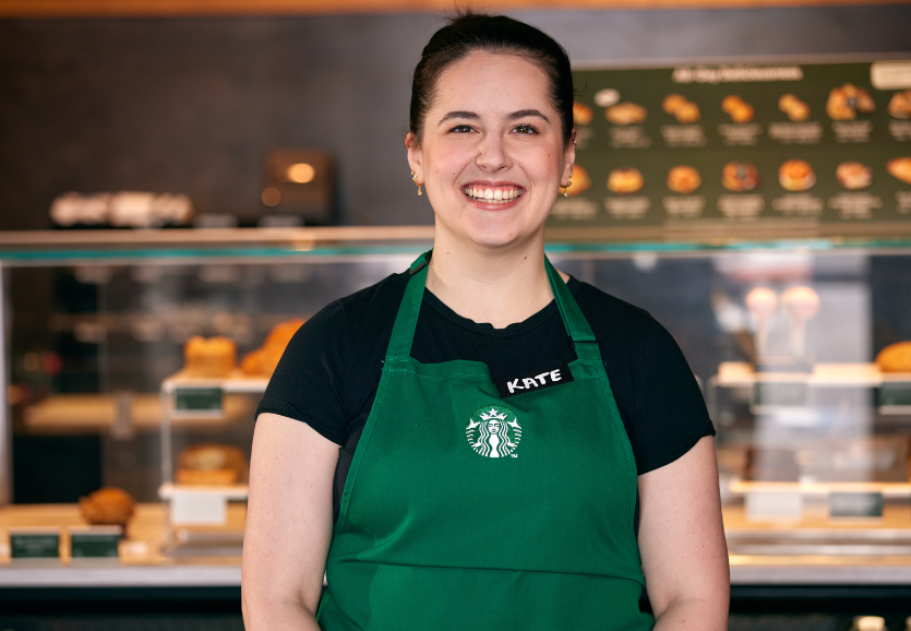 A Starbucks partner stands in a coffeehouse, wearing the green apron and smiles at the camera.