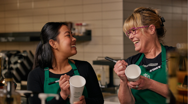 A Starbucks barista and her manager laugh together as they write on cups.