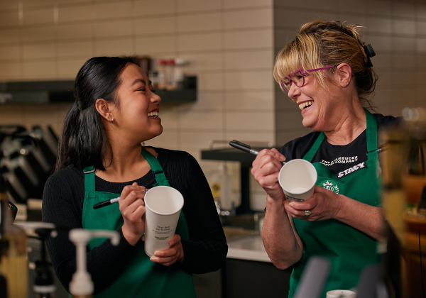 A Starbucks barista and her manager laugh together as they write on cups.