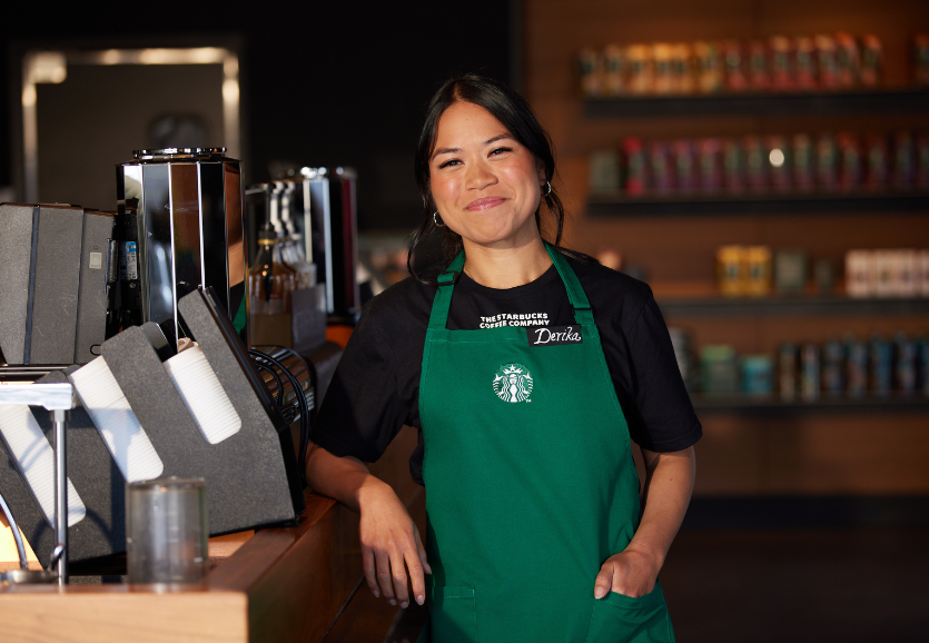 A Starbucks partner stands in a coffeehouse, wearing the green apron and smiles at the camera.