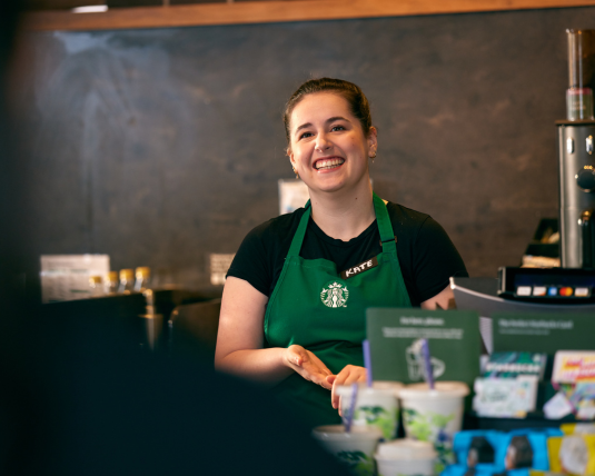 A Starbucks barista wearing a green apron, smiles at a customer.