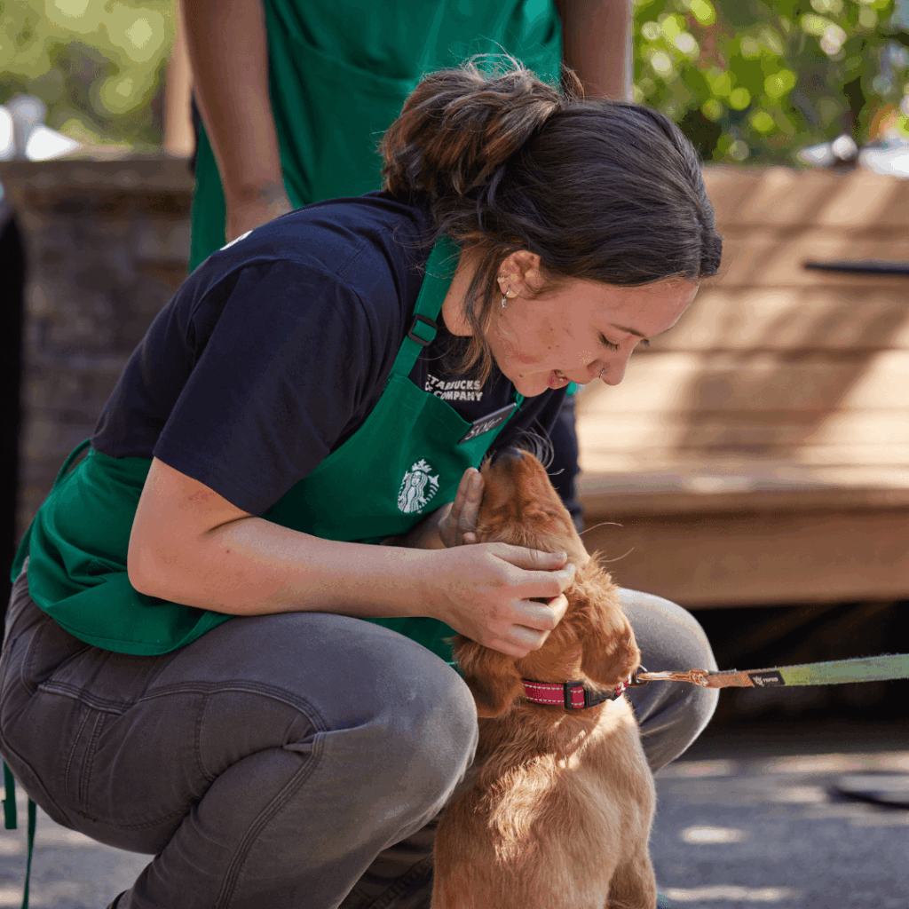A Starbucks barista crouches down to pet a dog in front of a Starbucks coffeehouse.