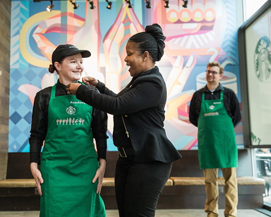 Deux personnes d’identités différentes sont debout côte à côte; l’une d’elles ajuste le col de l’autre qui porte un tablier vert Starbucks et lui sourit.